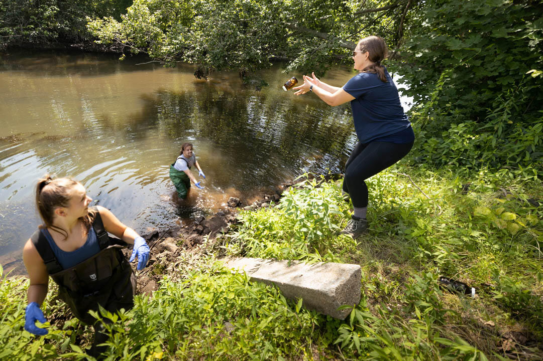 A student tossing a jar of water from inside of the river to a professor while another student climbs up the bank