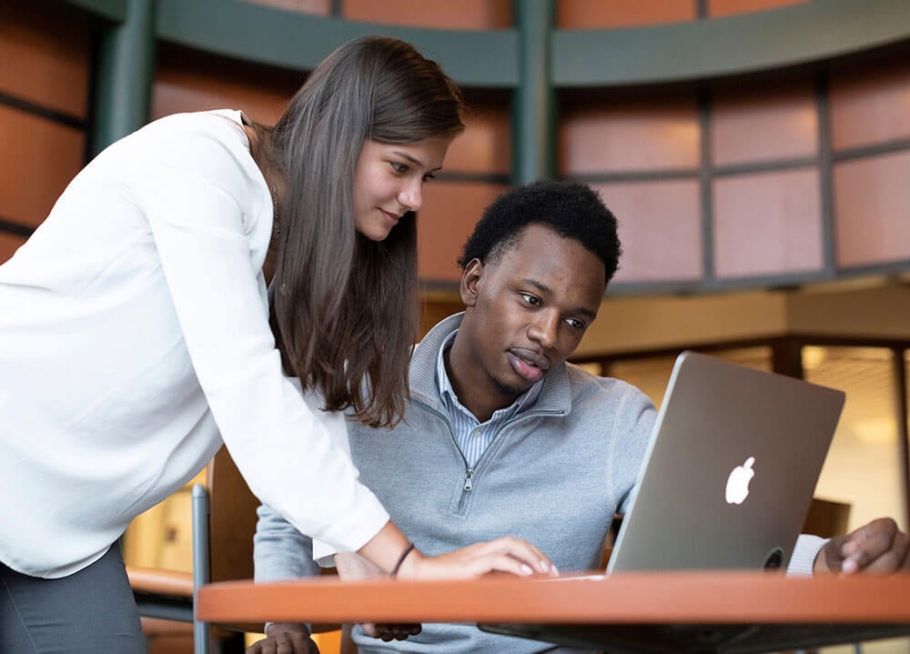Two students sit at a table and work on one open laptop