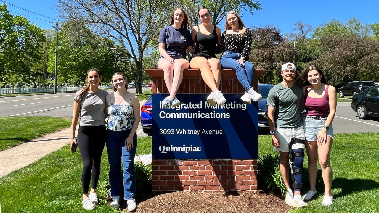 The Quinnipiac Today team stands in front of the Marketing and Communications office sign.