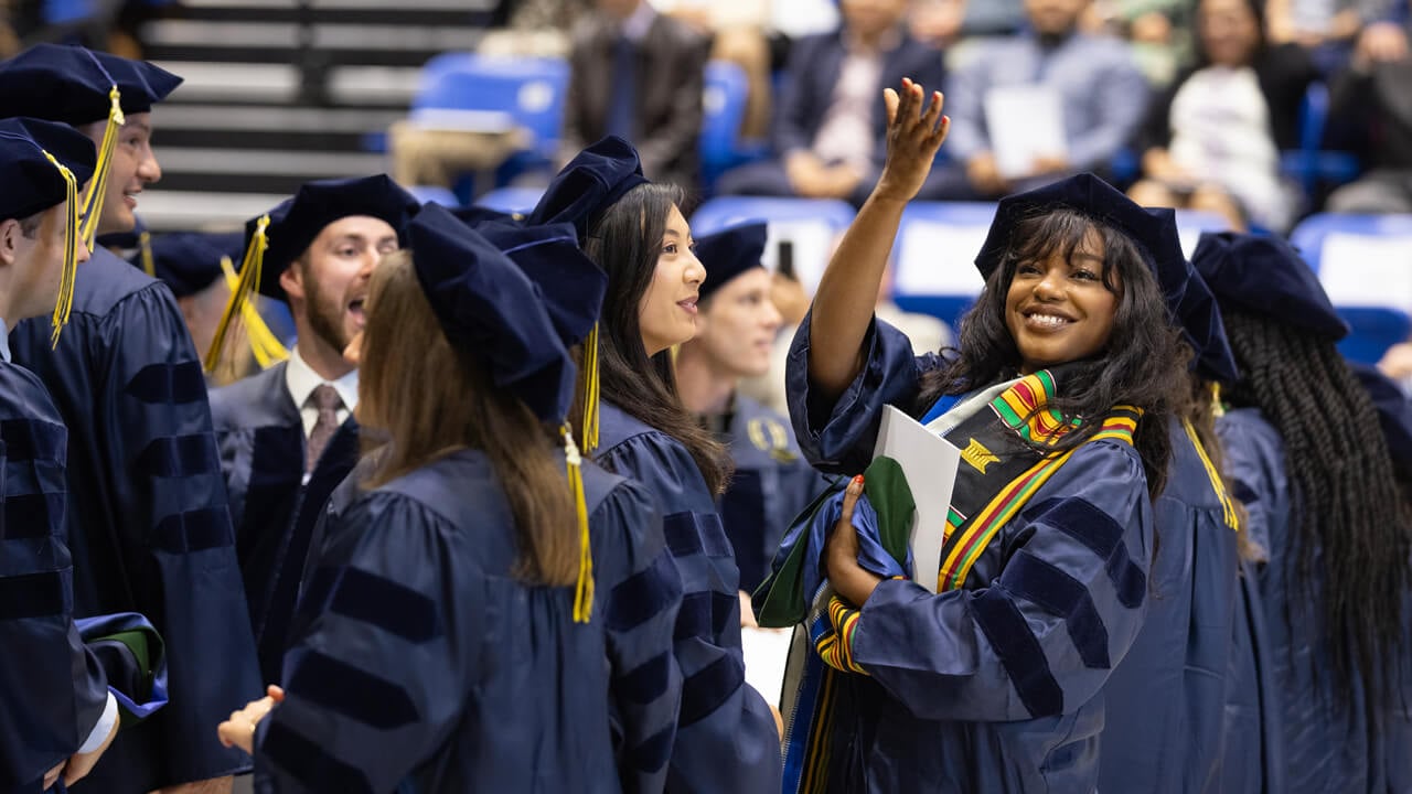 Medicine graduates cheer and wave to audience members while walking into Commencement