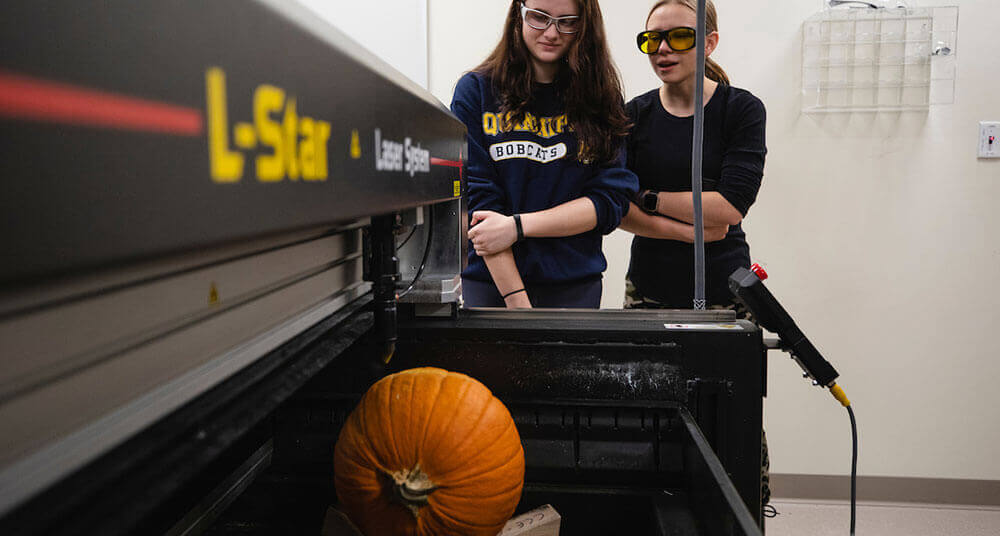 Two engineering students wearing safety goggles watch as their pumpkin is laser carved